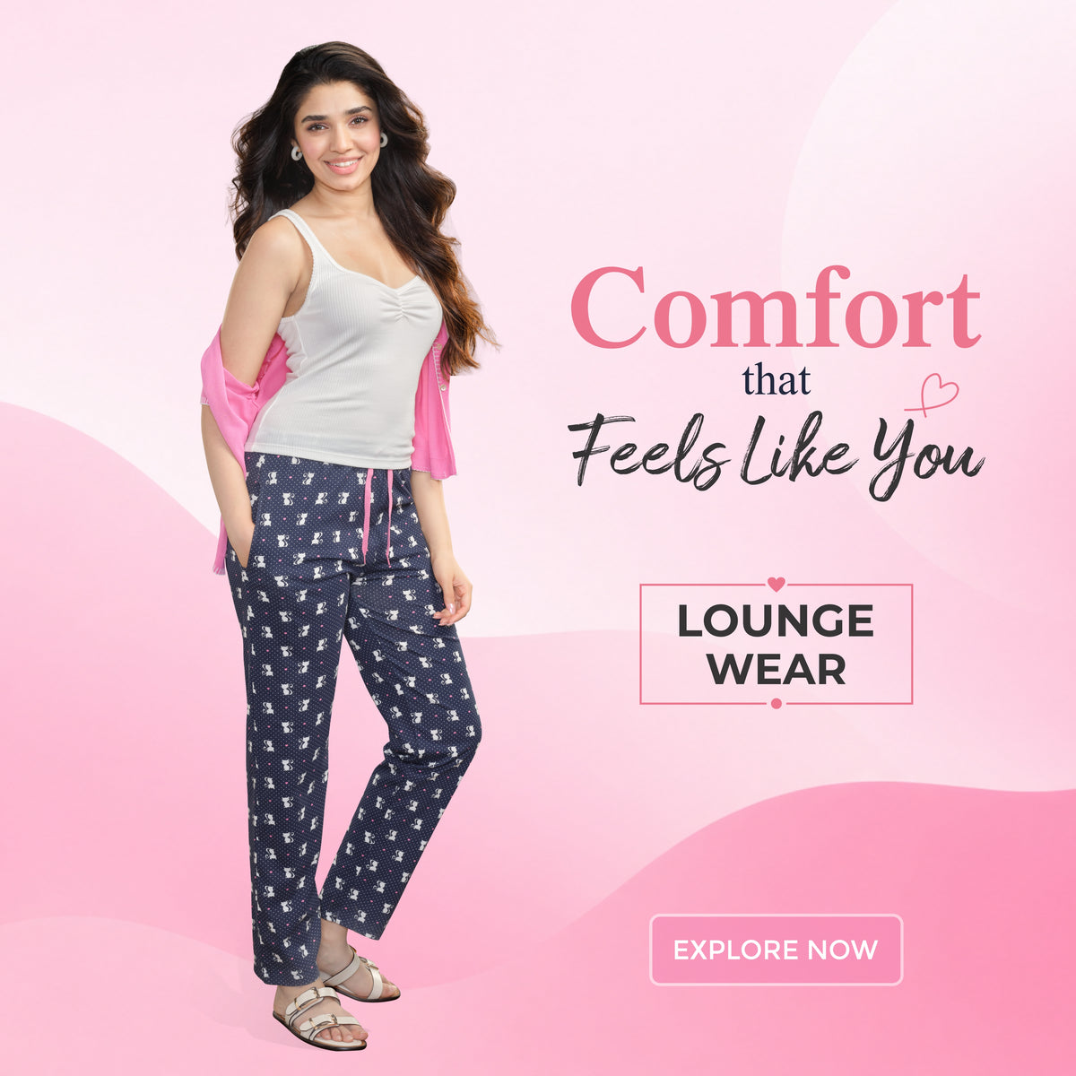 Actress Krithi Shetty modelling navy blue patterned lounge wear pants and a white tank top, with a pink cardigan draped off her shoulders, set against a pink graphic background.